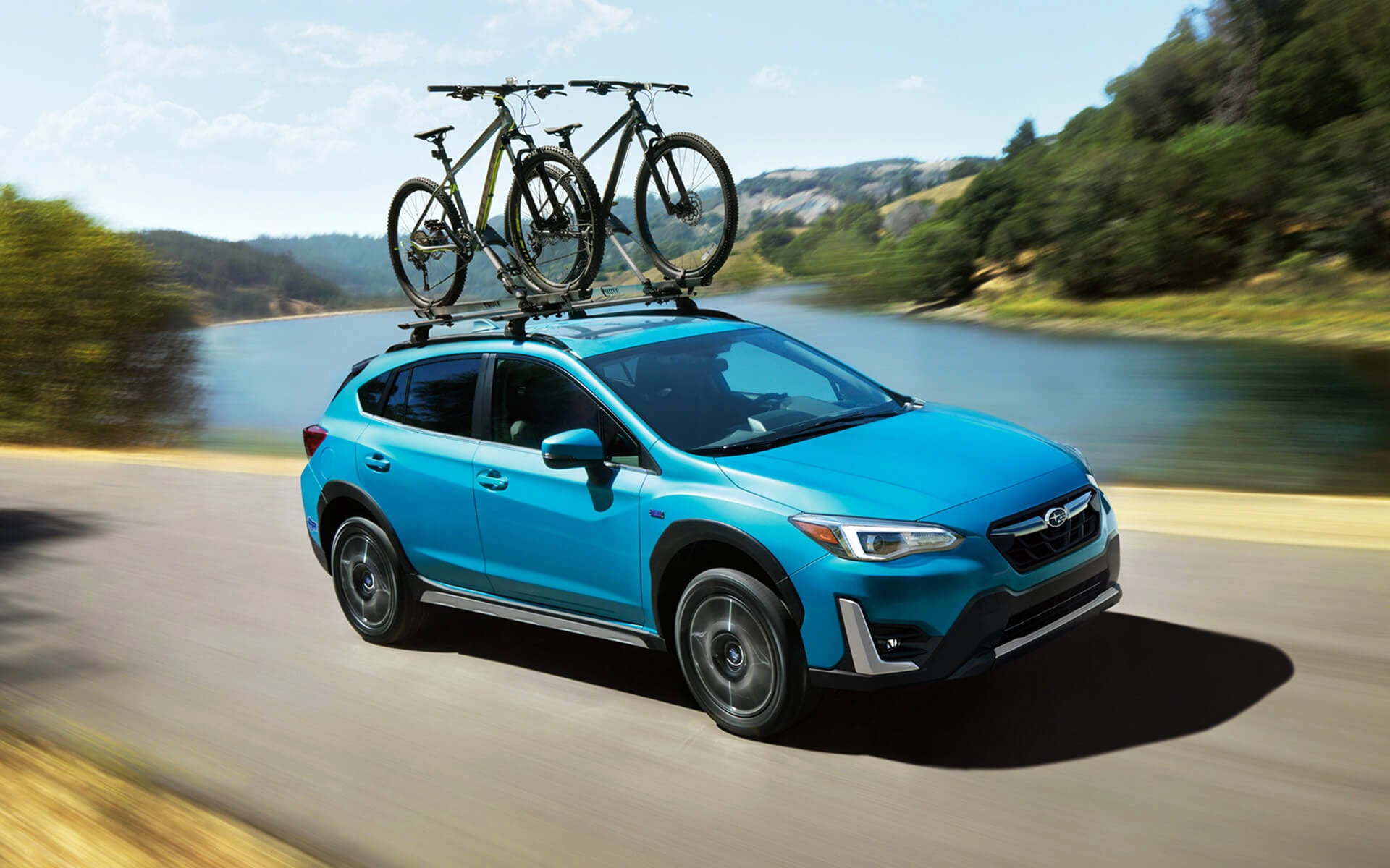 A blue Crosstrek Hybrid with two bicycles on its roof rack driving beside a river | All American Subaru of Old Bridge in Old Bridge NJ