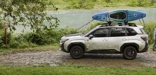 Front profile exterior static major of the Forester Wilderness in Crystal White Pearl with kayaks strapped to the roof rails and talent walking nearby.